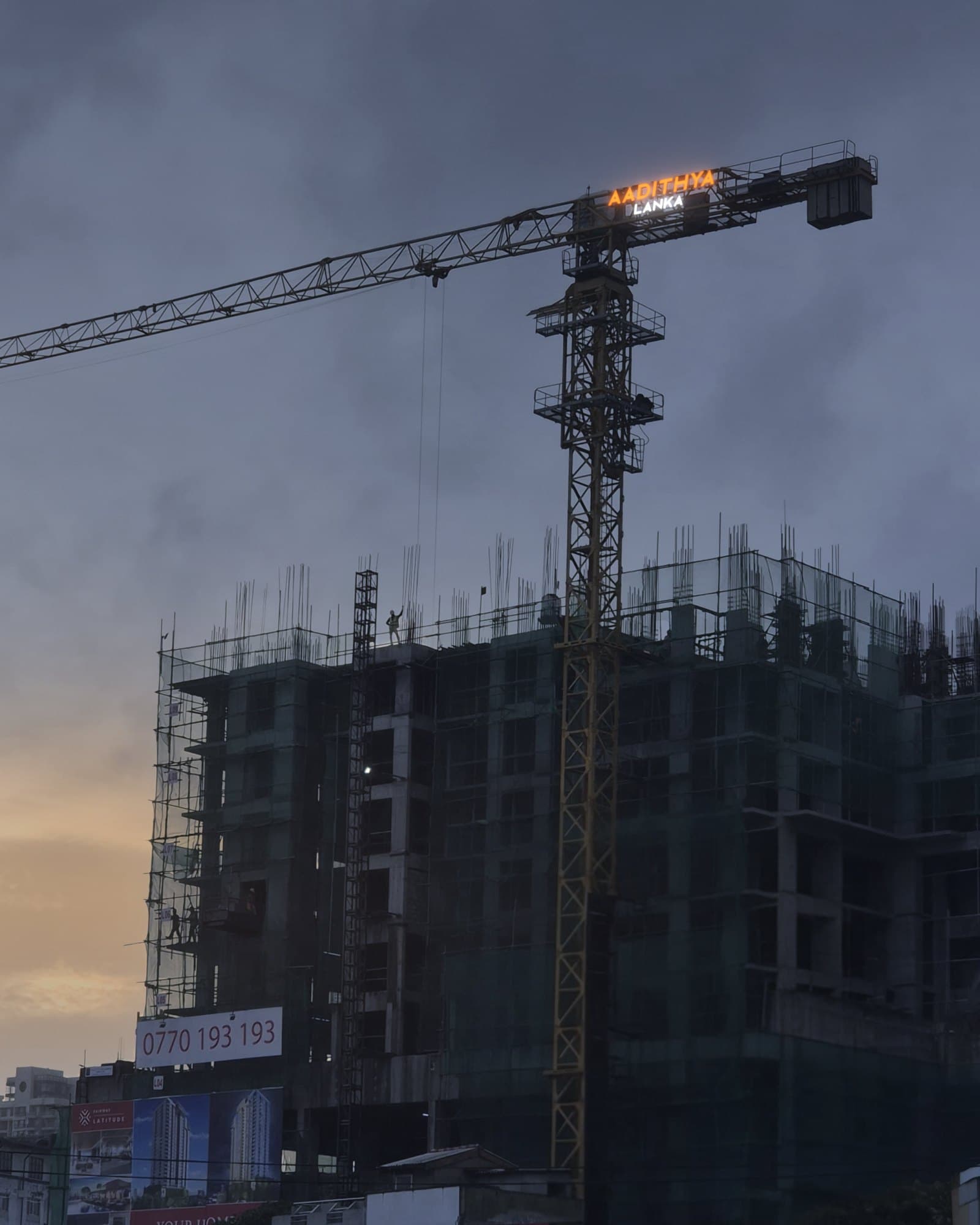 The Aadithya Lanka tower crane, its sign lit in the evening, over a residential tower under construction